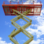 Scissor lift platform with hydraulic system at maximum height range painted in orange and beige colors, large construction machine, heavy industry, white clouds and blue sky on background